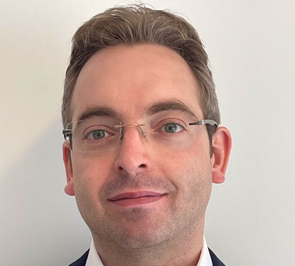 A man with short brown hair and glasses is wearing a dark suit jacket over a white shirt, standing in front of a plain light-colored background.